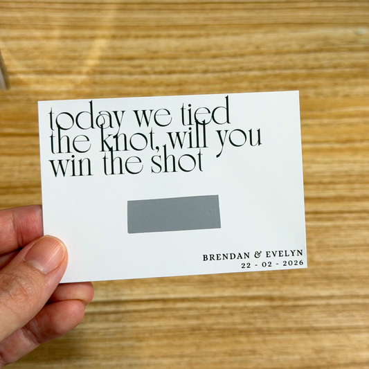 Card with text 'today we tied the knot, will you win the shot' held by a hand on a wooden surface