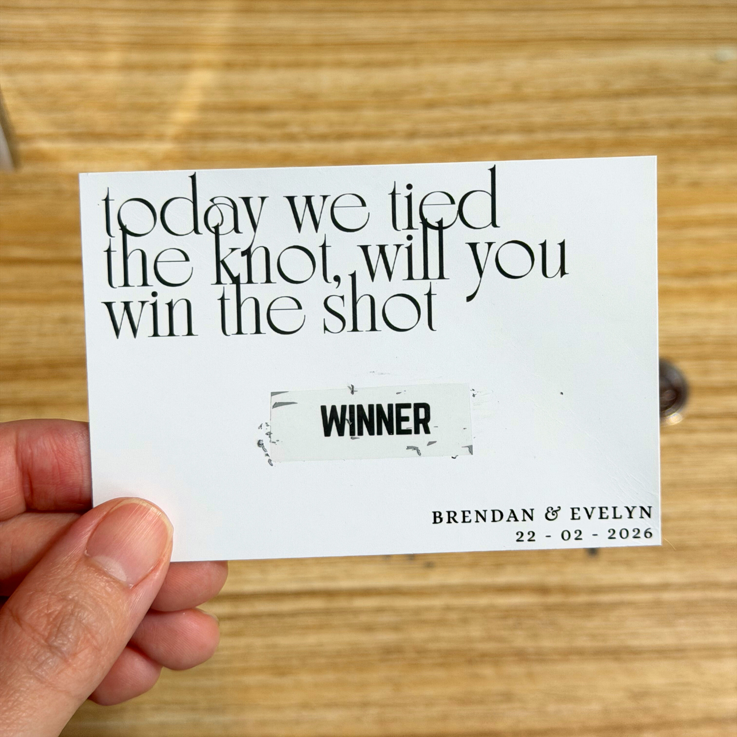 Card with 'today we tied the knot, will you win the shot' text held by a hand on a wooden surface.