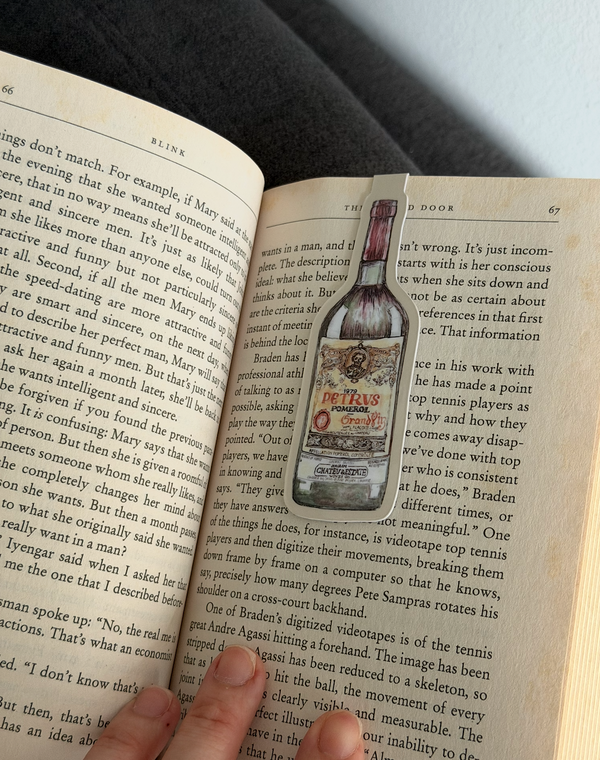 An open book with printed text and a printed illustration of a bottle labeled 'The Brimstone' overlaid on one page, with a hand holding the book open.