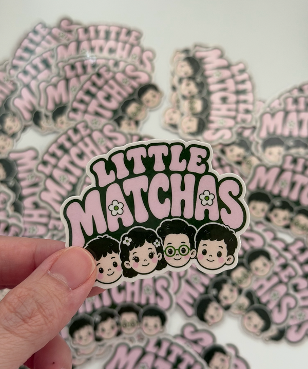 A hand holds a die-cut sticker that reads 'LITTLE MATCHAS' with cartoon girl and boy characters, while many similar stickers are scattered in the background.