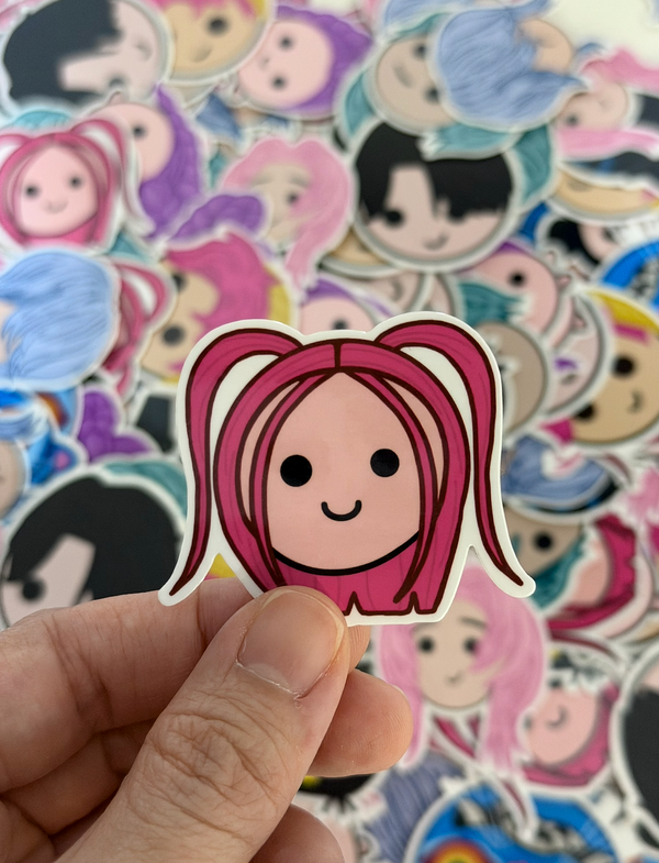 A hand holds a die-cut sticker featuring a cartoon girl with pink hair in pigtails, with a pile of colorful character stickers blurred in the background.