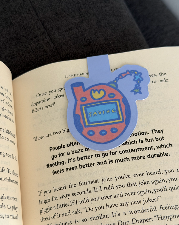 A die-cut paper bookmark shaped like a Tamagotchi toy, placed on an open book page. The bookmark is light blue with a red body, yellow smiley face, and a blue screen that reads 'SAVING'.