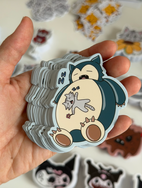 A hand holding a stack of die-cut stickers featuring a cartoon sleeping creature holding a small gray cat. The stickers have a light blue outline, with the character colored in dark teal, cream, and brown, and the cat in light gray.