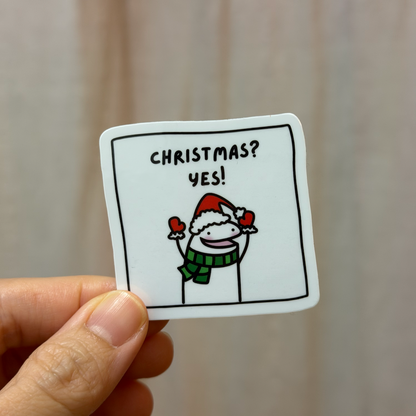 Hand holding a sticker with a snowman and text 'Christmas? Yes!' against a plain background
